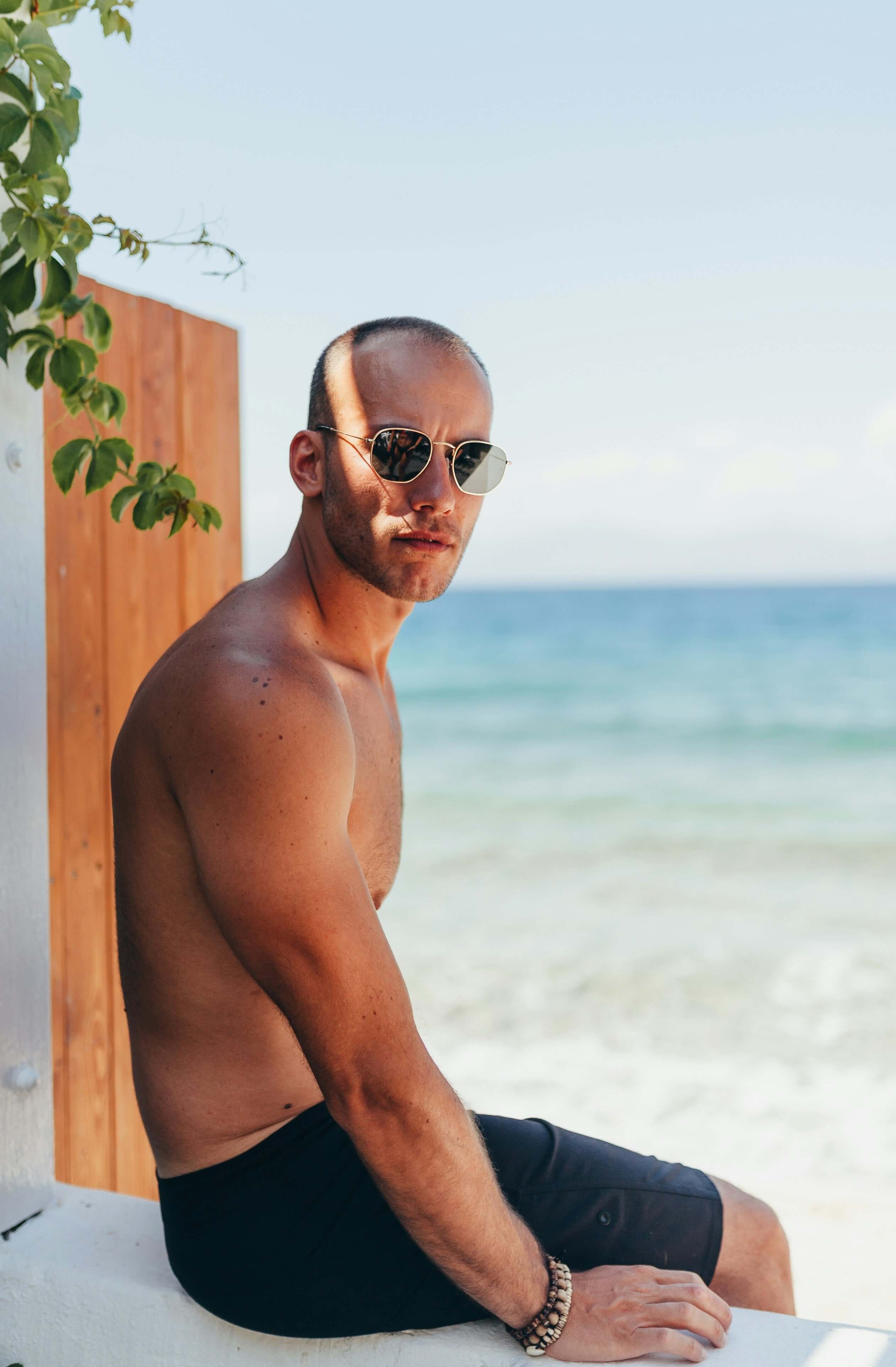 A man in sunglasses sits casually on a stone wall, enjoying the outdoors at the beach.