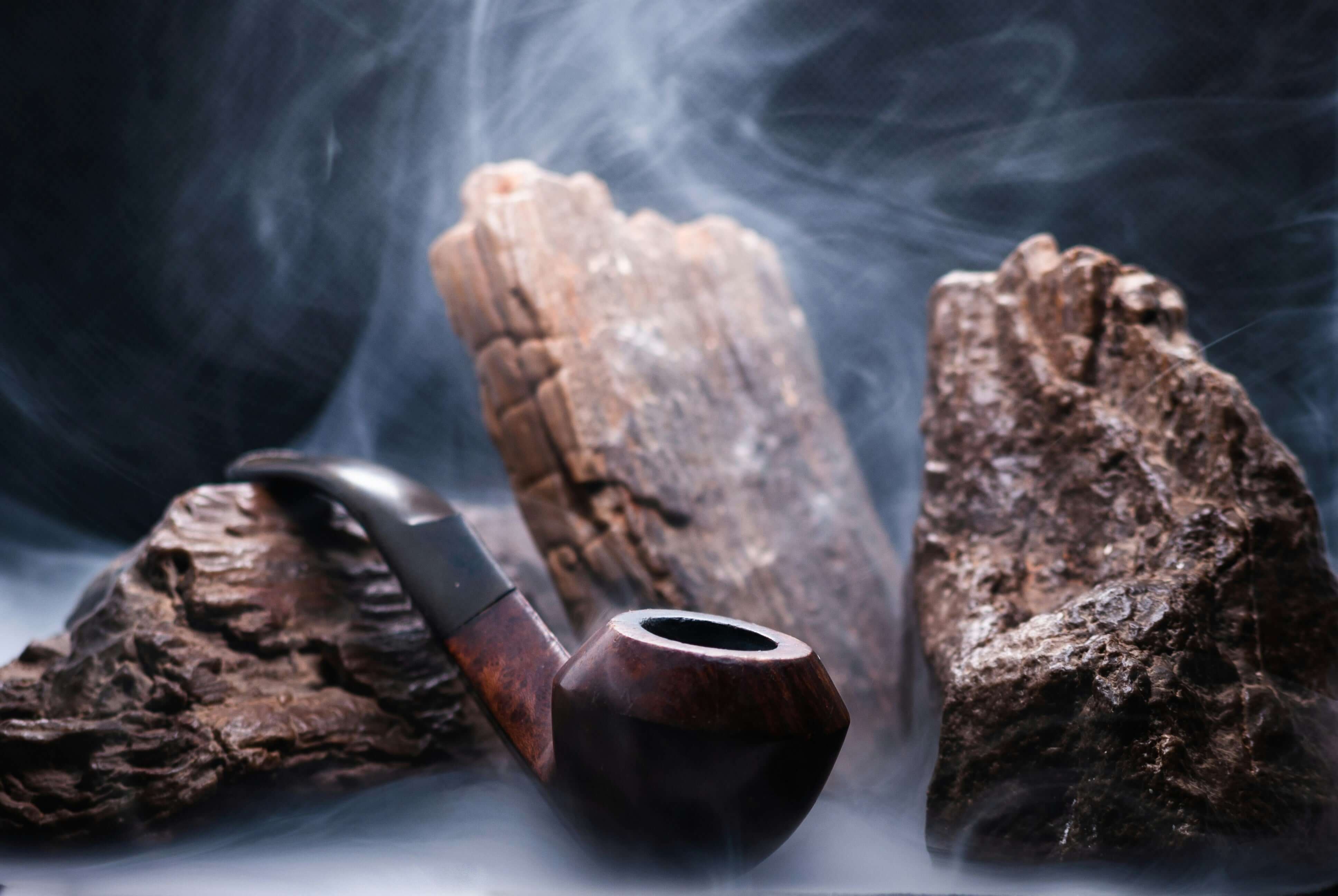 A pipe rests beside a rock, with wisps of smoke rising from the rock's surface, creating a mysterious atmosphere.