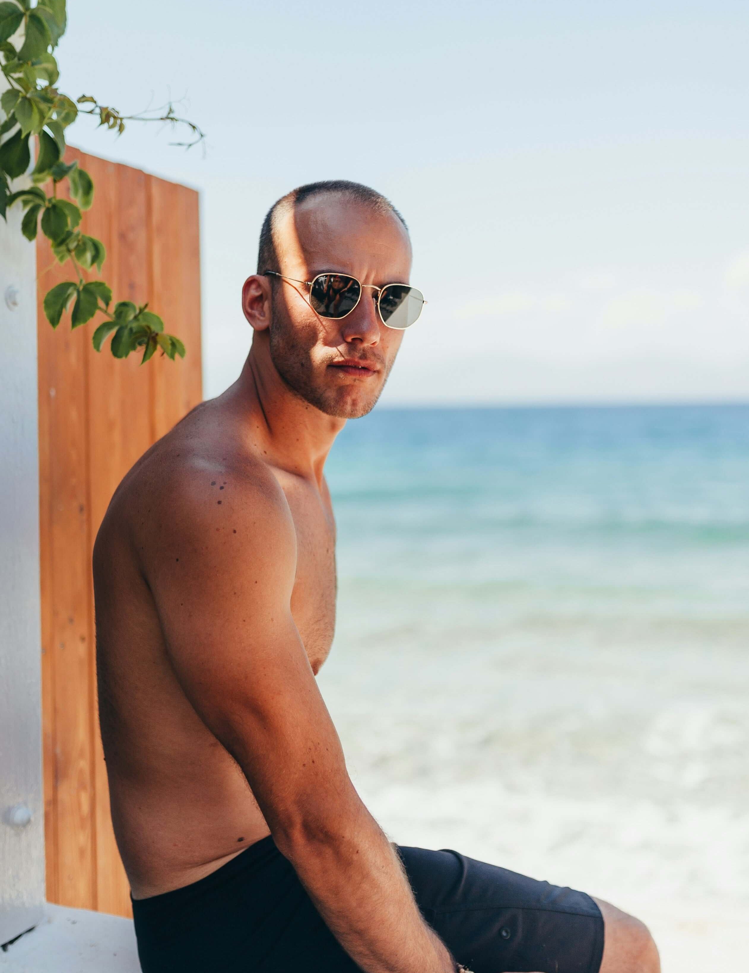 A man in sunglasses sits casually on a stone wall, enjoying the outdoors at the beach.