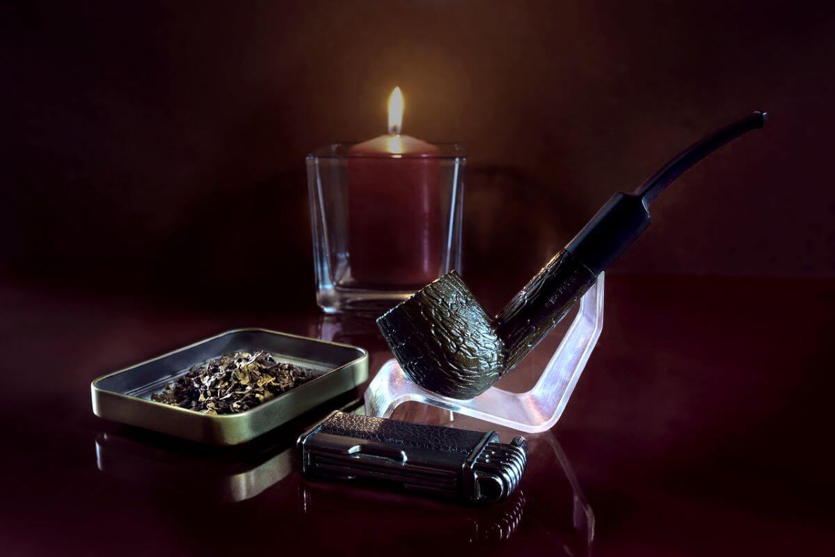A pipe rests beside a lit candle and a glass of bourbon on a wooden table, creating a cozy atmosphere.