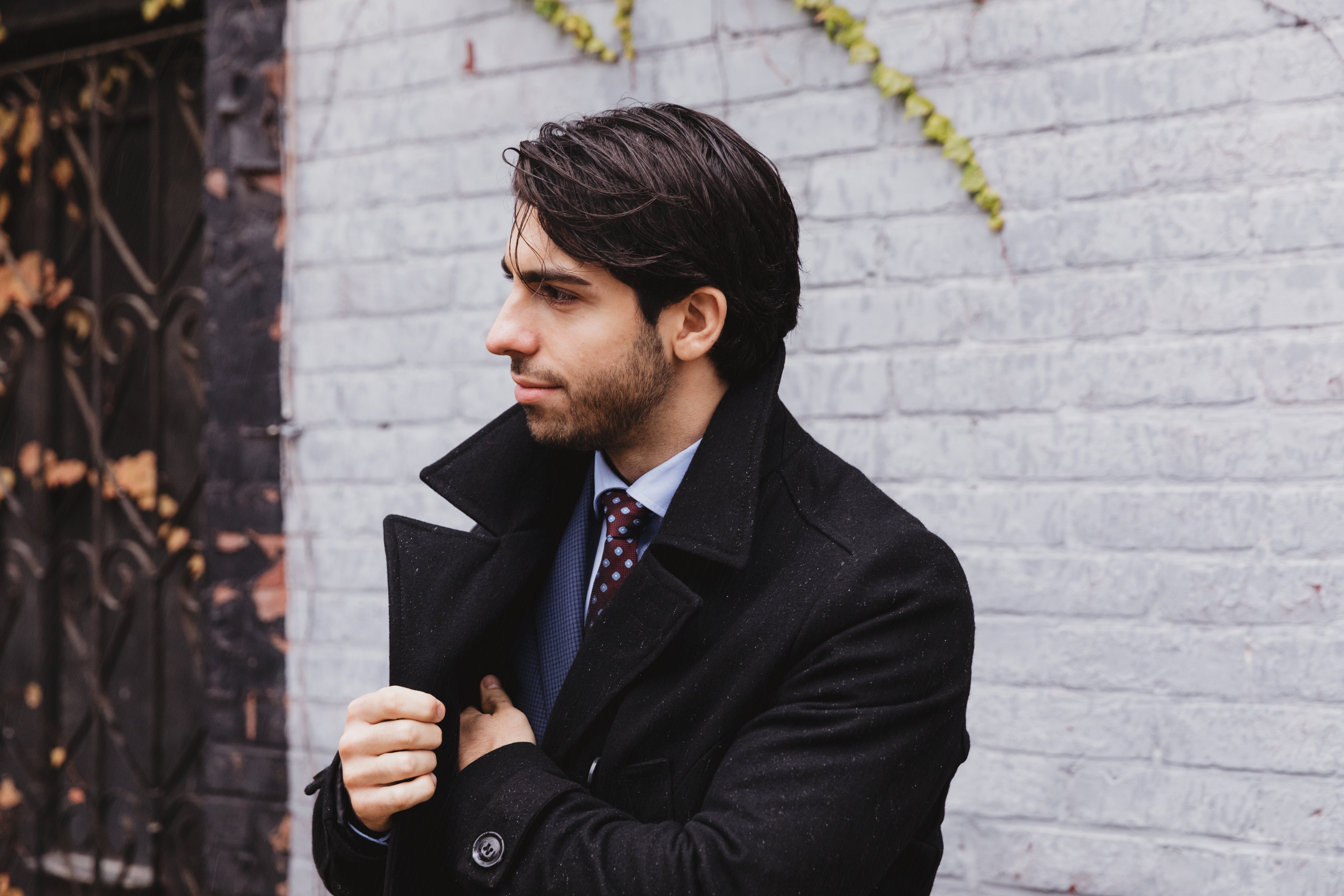 A man wearing a dark jacket stands casually, with a neutral expression on his face.
